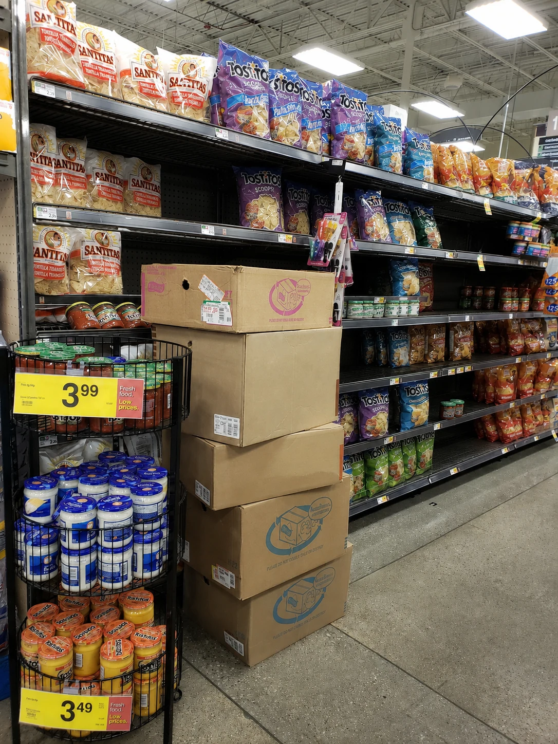 A supermarket snack aisle with a stack of unopened Frito-Lay delivery cases on the floor beside long runs of empty shelf facings.